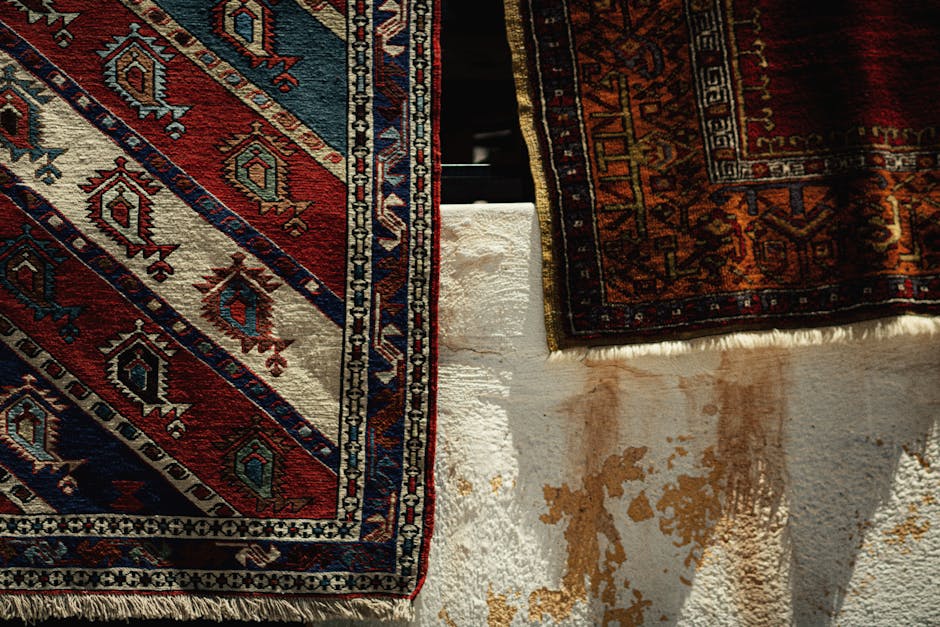 Close-up view of two traditional Persian-style rugs with intricate geometric and floral patterns in shades of red, blue, beige, and orange, draped over a white textured surface with visible stains and