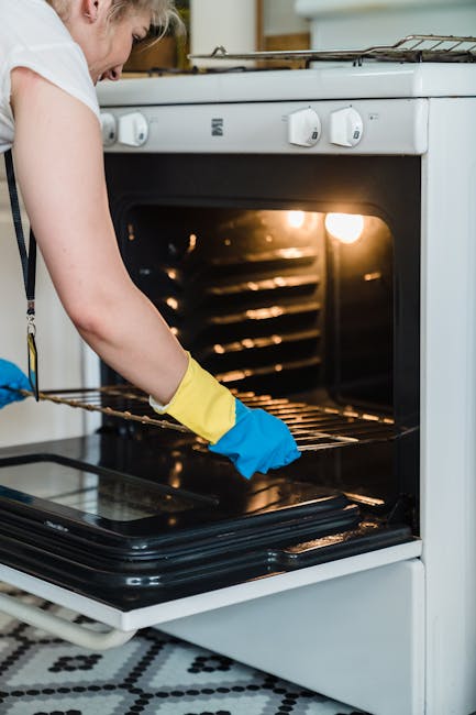 A person wearing yellow and blue rubber gloves is cleaning the interior of a black oven at home, using a pink cloth to wipe the oven surface. The oven is part of a white gas stove with visible burners